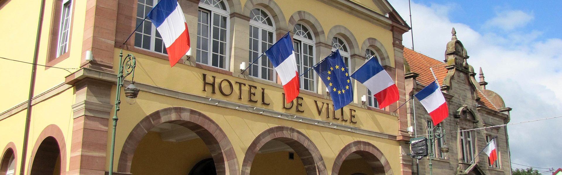 Vue de la mairie de Brumath avec ses arcades en grès rose, sa façade jaune restaurée, des drapeaux officiels et des jardinières de fleurs en fleurs.