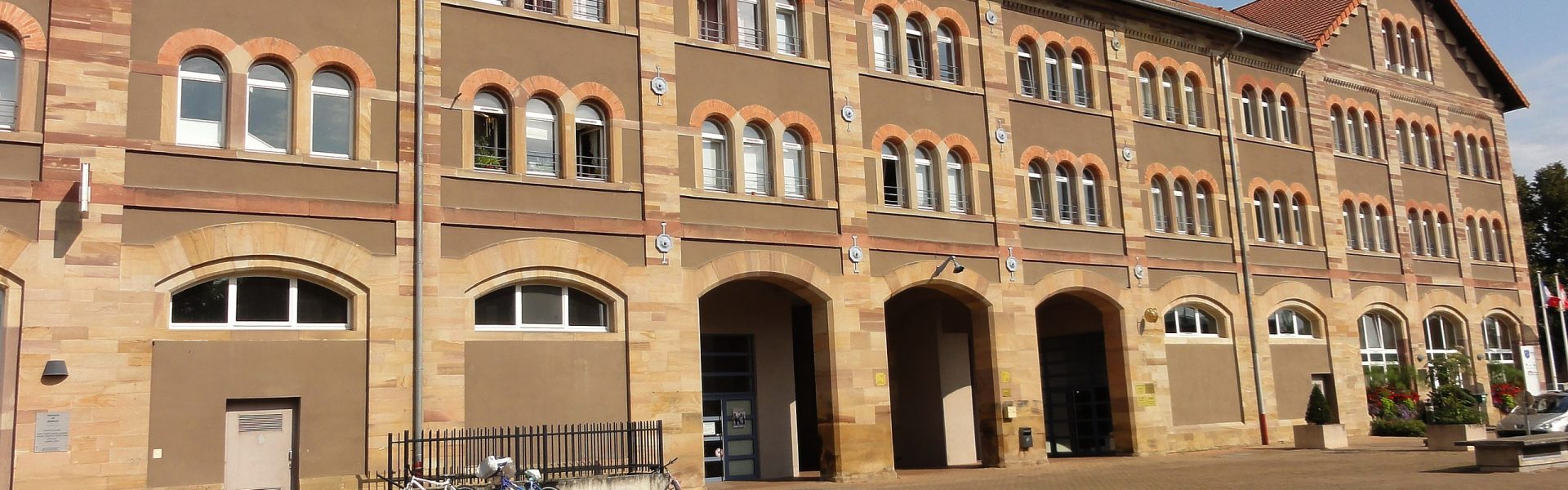 Façade rénovée de l'ancienne manufacture de tabac à Benfeld avec crépi brun, arches en grès et multiples fenêtres symétriques.