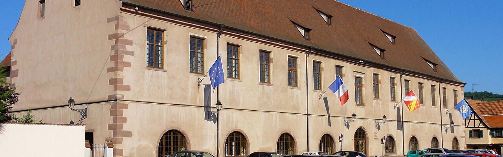 Vue d'ensemble de l'Hôtel de la Monnaie à Molsheim : grand bâtiment historique alsacien avec murs en crépi beige, fenêtres et angles en grès rose, sous un ciel bleu.