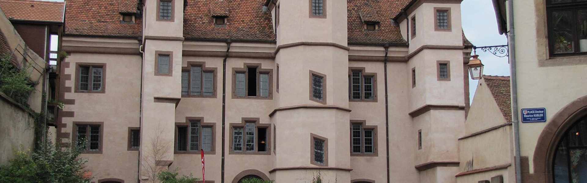 Bâtiment historique alsacien aux murs beiges, doté d'une tour d'escalier, de fenêtres sculptées et d'un grand toit de tuiles rouges.