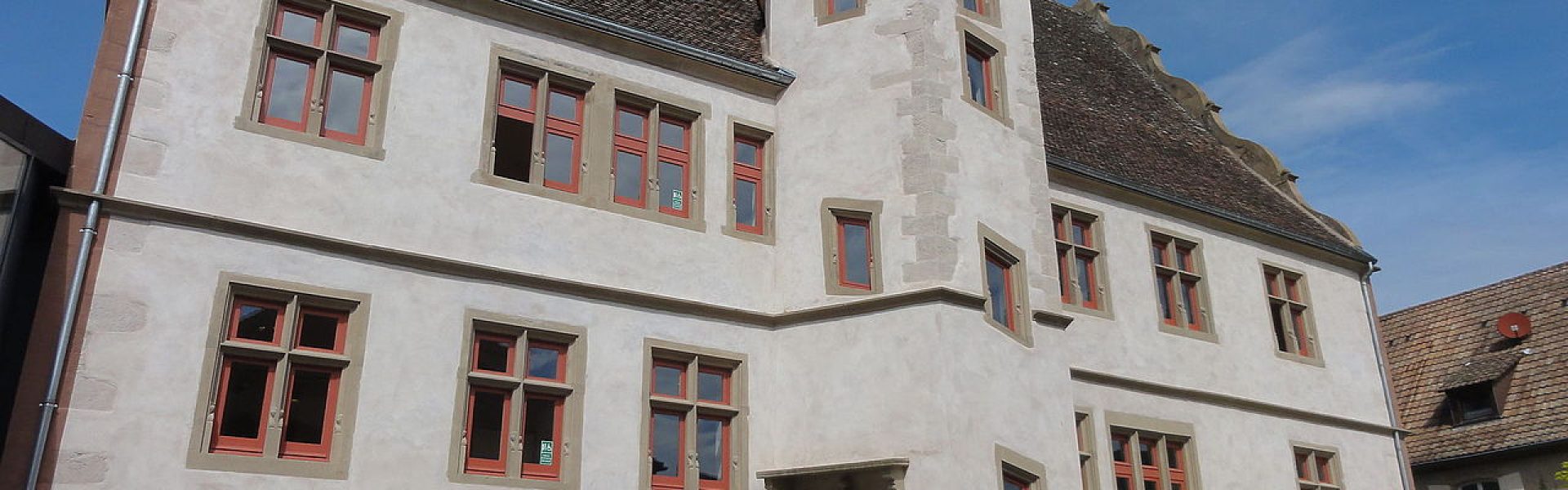 Grand bâtiment historique à Andlau avec enduit clair, tour seigneuriale, fenêtres à meneaux et portes rouges.
