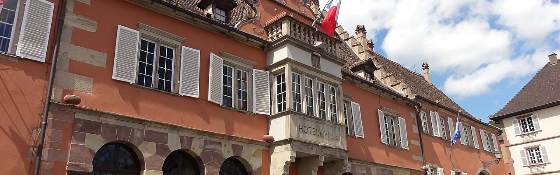 Façade monumentale de l'Hôtel de Ville de Barr avec oriel sculpté, arcades en grès rose et volets blancs sous un ciel bleu.