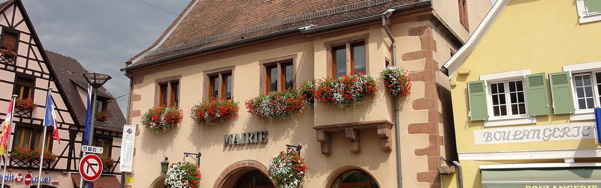 Façade restaurée de la mairie de Châtenois avec pierres de taille en grès rouge, crépi ocre, horloge et balcons fleuris sous un ciel nuageux.