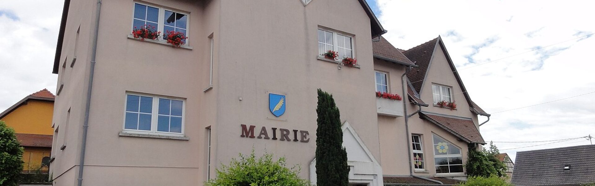 Façade de la mairie de Huttendorf en crépi rose pâle avec lettrage moderne, fleurs aux fenêtres et ciel nuageux.