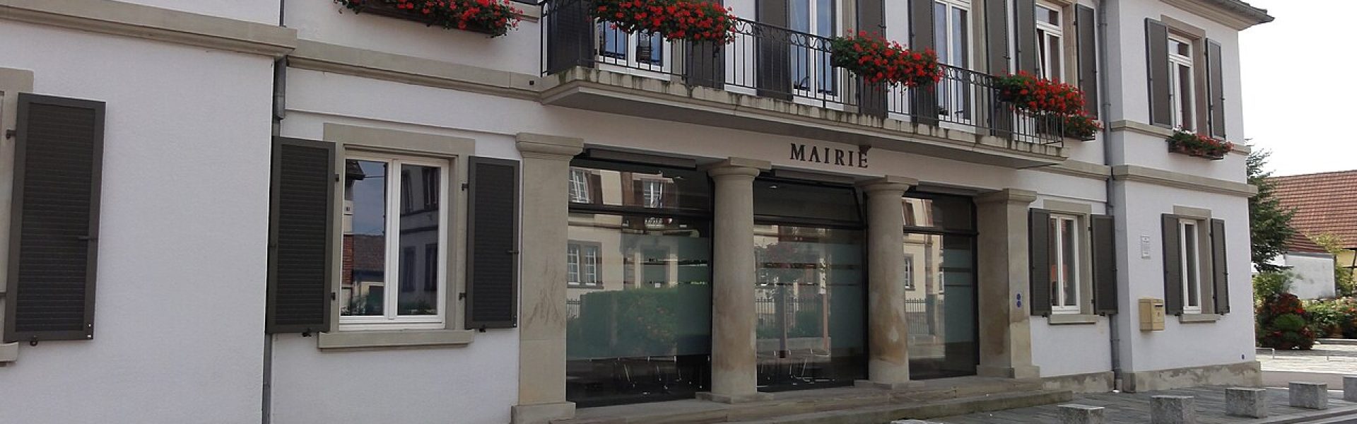 Bâtiment de la mairie de Huttenheim avec une façade blanche rénovée, des volets sombres et des jardinières fleuries de géraniums rouges.