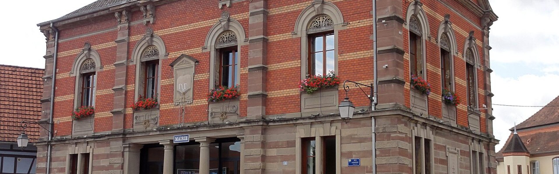 Façade de la mairie de Kertzfeld en briques rouges et jaunes avec soubassement en grès des Vosges, fenêtres fleuries et colonnes à l'entrée.