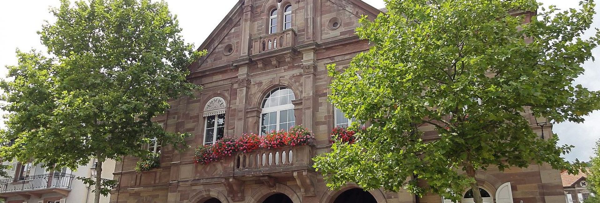 Imposante mairie de Kintzheim construite en pierre de grès rouge avec trois grandes arcades à l'entrée et deux arbres au premier plan.
