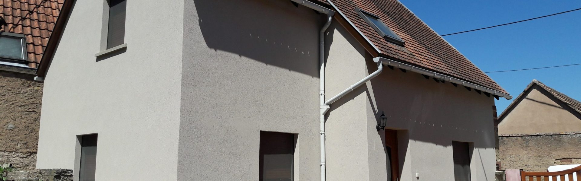 Vue d'une maison rénovée avec un crépi extérieur beige clair, fenêtres sombres et toiture en tuiles sous un ciel bleu.