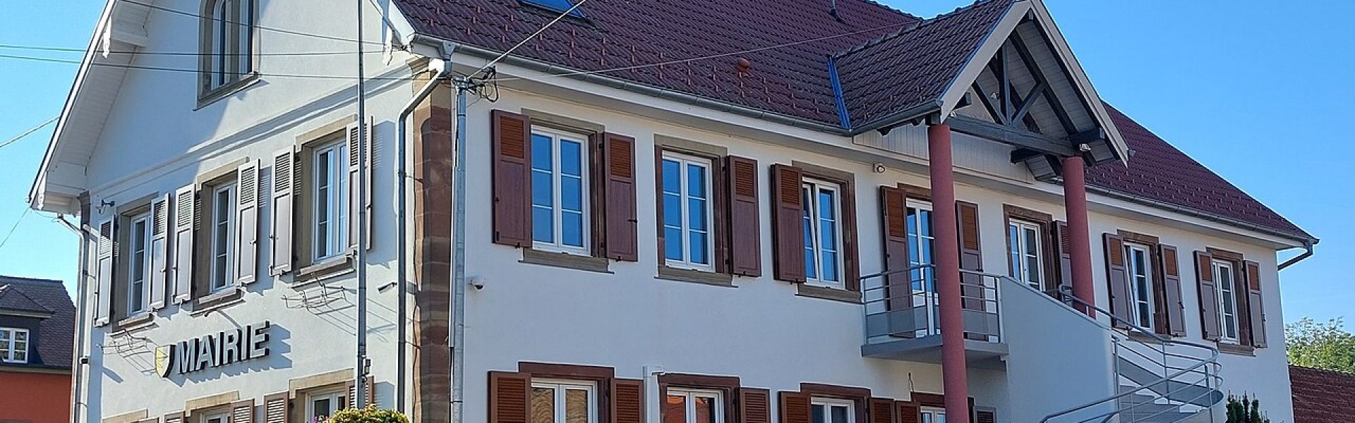 Vue de la mairie de Bilwisheim : bâtiment alsacien à façade blanche, volets marron, escalier extérieur moderne et nombreuses fleurs colorées sous un ciel bleu.
