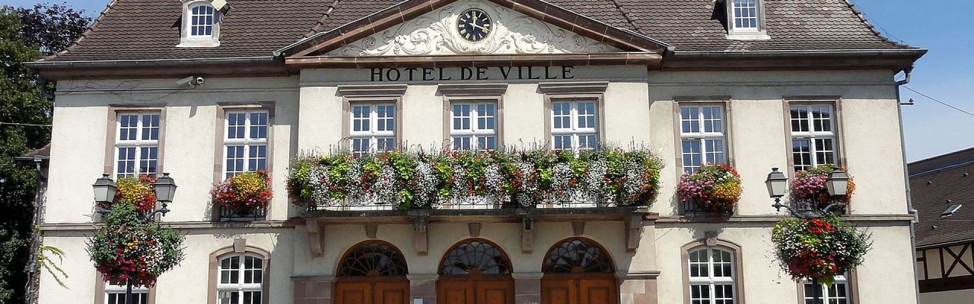 Façade de l'Hôtel de Ville d'Erstein avec balcons fleuris, horloge et drapeau français sous un ciel bleu.