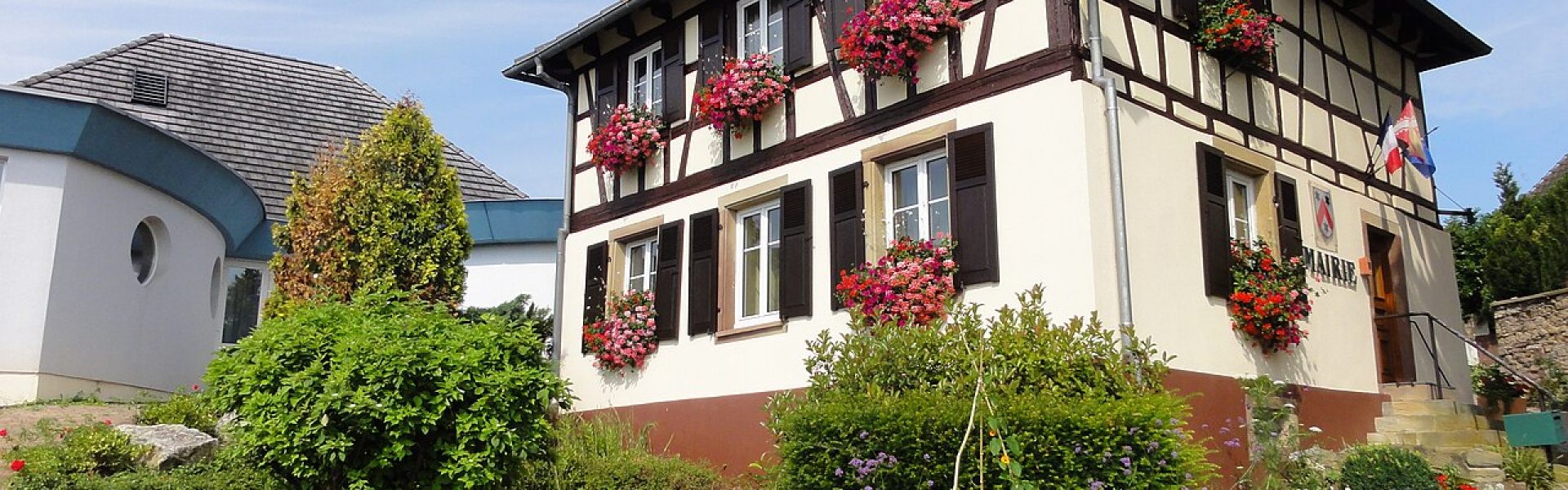 Mairie de Durningen, bâtiment traditionnel alsacien avec murs blancs, poutres apparentes brunes et jardinières de fleurs rouges.