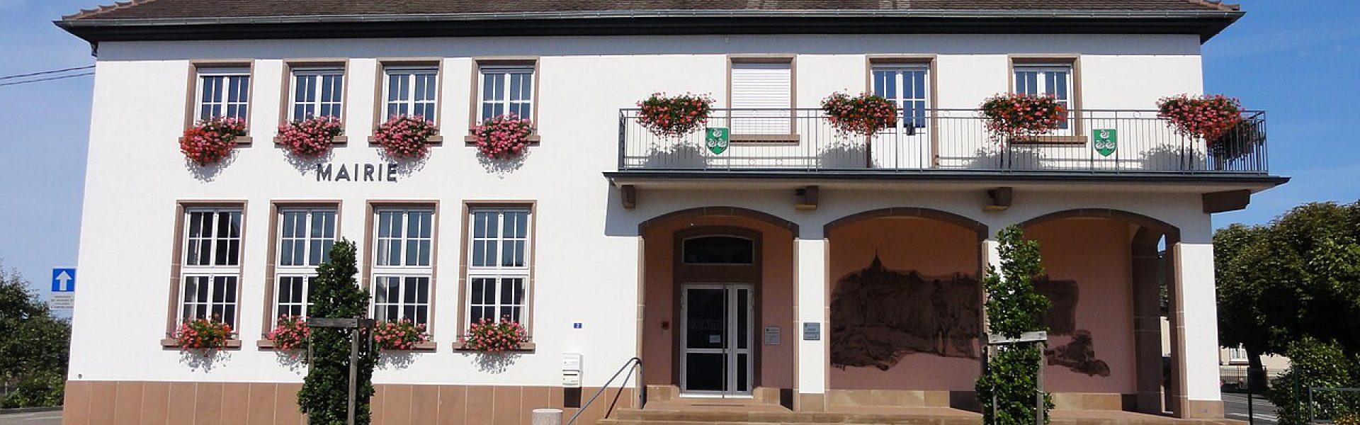 Façade de la mairie de Sessenheim avec crépi blanc, fenêtres fleuries de géraniums et balcon sous un ciel bleu dégagé.